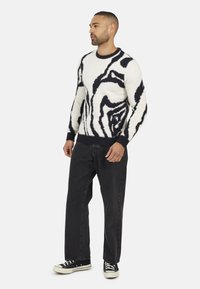 Le pull en tricot présente une base blanche avec des motifs abstraits noirs, un col rond et des poignets côtelés. Associé à un jean foncé et des baskets noires.