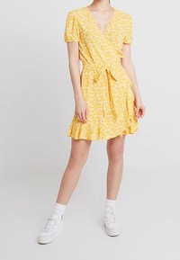 Robe portefeuille jaune à motifs floraux avec des manches courtes bouffantes, un décolleté en V, une taille à nouer et un ourlet ondulé. Portée avec des baskets blanches et des chaussettes mi-mollet.