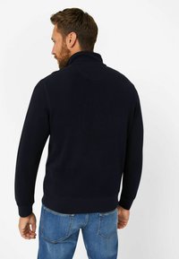 Marineblauer Fleece-Pullover mit hohem Kragen, gerippten Bündchen und Saum. Weiche Textur mit einem schlichten Rückendesign, kombiniert mit hellblauen Jeans.