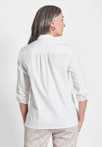 Weißes Hemd mit hochgekrempelten Ärmeln und einem gefältelten Rücken. Glatte Textur, klassischer Kragen, kombiniert mit gemusterten hellen Hosen.