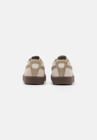 Sneaker in suede beige con suola in gomma marrone. Le caratteristiche includono un colletto imbottito, strisce laterali a contrasto e un design del tacco arrotondato.