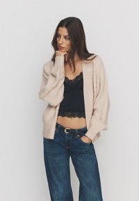 Vrouw die een beige vest draagt over een zwart kanten cropped top en een blauwe spijkerbroek, staand met één hand in de zak en de andere hand die haar kin aanraakt.