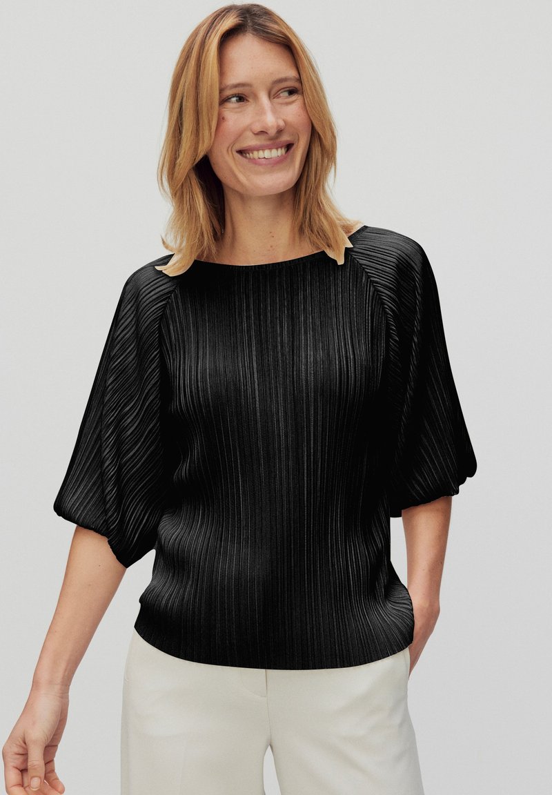 Sort plisseret top med rund halsudskæring og korte, puffede ærmer. Har en subtil tekstur og en glat finish, parret med hvide bukser.