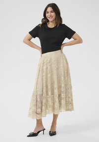 Gonna midi beige in pizzo con un motivo floreale, abbinata a una t-shirt nera e mule con tacco neri. La gonna presenta una vita aderente e un design fluido.
