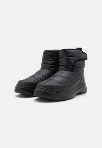 Botas de estilo puff negra con textura acolchada, suela de goma gruesa y un tirador. Presentan un branding minimalista en el costado.