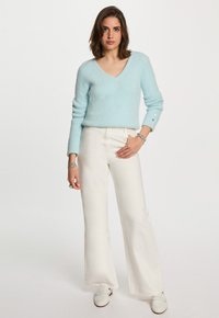 Femme portant un pull V en maille bleu clair, un pantalon large blanc, des mocassins blancs et des bracelets en argent, se tenant devant un fond uni.