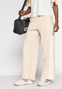 Bézs széles szárú nadrág fehér Adidas csíkokkal és cipzárokkal a szárvégeken, fehér sportcipővel és fekete utazótáskával párosítva.