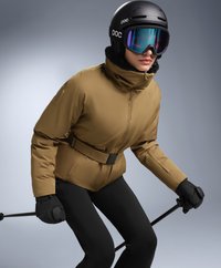 Brown insulated ski jacket with a belt, black ski pants, black gloves, and a black helmet. Tinted goggles add color contrast.