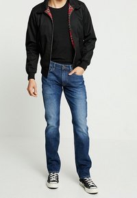 Chaqueta negra con cremallera y forro de cuadros rojos, camiseta negra ajustada, vaqueros azules y zapatillas negras con suelas blancas. Conjunto informal.