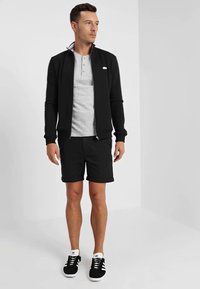 Schwarze Zip-Jacke, graues Henley-Shirt, schwarze Shorts und schwarze Sneaker mit weißen Akzenten. Hat eine schmale Passform und strukturierten Stoff.