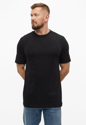 Mężczyzna z krótkimi włosami i zarostem, ubrany w zwykły czarny t-shirt i niebieskie dżinsy, stoi z rękami za plecami, patrząc w lewo.