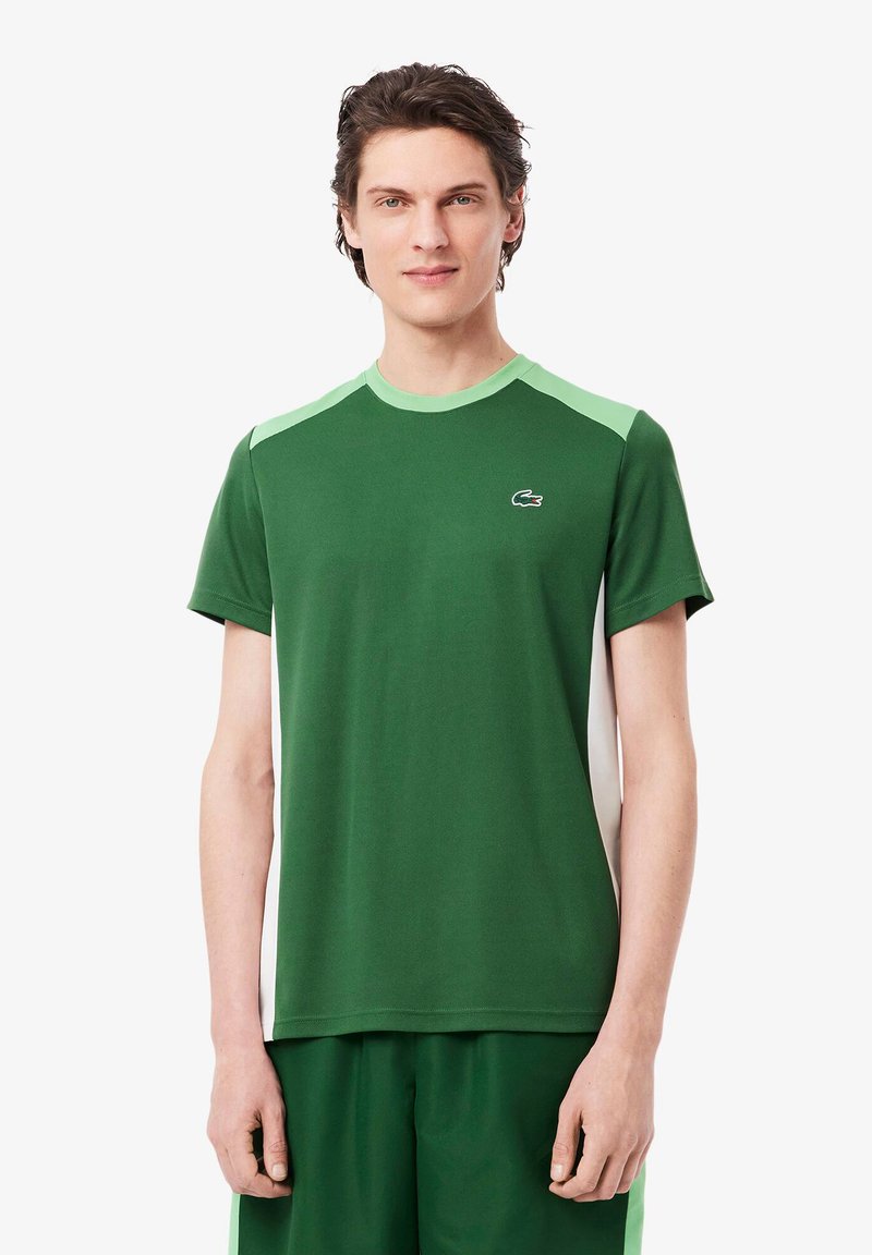 Tricou sportiv verde, confecționat dintr-un material moale, cu guler rotund și accente de umăr de culoare verde deschis în contrast. Logo-ul Lacoste pe piept.