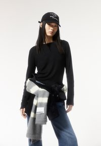 Schwarzes Langarmshirt, blaue Jeans, grau-weiß gestreifter Schal um die Taille gebunden, schwarze Mütze mit weißen Schriftzug, glatte Textur.