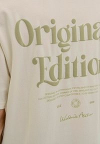 Kremos spalvos marškinėliai su dideliu, įspaustu alyvų žaliu tekstu „Original Edition“. Minkšta tekstūra ir laisvas kirpimas. Detalės apima apvalų logotipą ir papildomą tekstą.