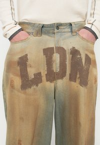 Τζιν παντελόνια με ξεθωριασμένο, πολύχρωμο φινίρισμα. Μεγάλα καφέ γράμματα "LDN" στο πίσω μέρος, με φθορές και διακριτικές παραλλαγές υφής.