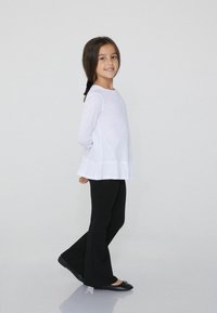 Weißes Langarmoberteil mit Rüsche am Saum, kombiniert mit schwarzen, weit geschnittenen Hosen und schwarzen Ballettschuhen. Model steht seitlich mit geflochtenem Haar.