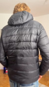 Casaco puffer preto com capuz ajustado, textura acolchoada e detalhes de costura visíveis, usado sobre jeans azuis.