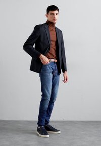 Blazer preto ajustado sobre um suéter de gola alta castanho, combinado com jeans azuis e ténis escuros. Texturas suaves; silhueta ajustada.