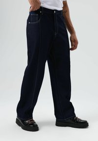 Donkere denim broek met rechte pijpen, lichte stiknaden, zakken aan de voorkant en een knoopsluiting. Zwarte schoenen met meerkleurige veters.
