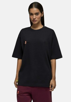 Femme portant un t-shirt noir oversize avec une petite poche poitrine et un pantalon rouge foncé avec des détails de fermeture éclair orange.