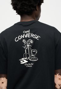 Μαύρο μπλουζάκι με κοντό μανίκι που διαθέτει λευκό γραφικό σχέδιο με τριαντάφυλλ α, φλιτζάνια καφέ και το κείμενο "Café Converse" και "Φρεσκοφτιαγμένο."