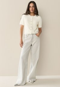 Cardigan bianco e soffice a maniche corte con chiusura a bottoni, abbinato a jeans grigio chiaro a gamba larga con cuciture decorative sulle tasche.