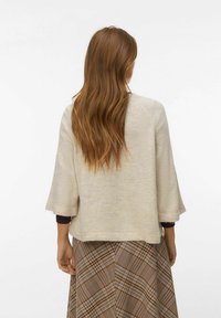 Femme aux longs cheveux bruns ondulés portant un haut beige clair à manches larges et une jupe marron à carreaux, debout de dos.