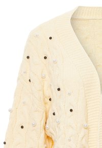 Krämbelagd kabelstickad cardigan som har strukturerade mönster och dekorationer av vita och svarta pärlor. V-ringad design med ribbade kanter.