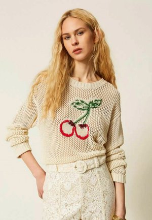 Donna bionda che indossa un maglione lavorato a maglia color crema con ricamo di ciliegie rosse e pantaloni in pizzo bianchi con cintura abbinata, in piedi davanti a uno sfondo semplice.