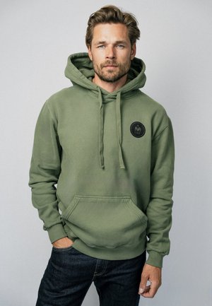 Man met lichte baard, dragend een groen capuchontrui met voorzak en een zwart rond logo op de borst, staand tegen een effen achtergrond.