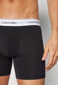 Melni kokvilnas šorti bokseru veidā ar baltu elastīgu jostu, uz kuras redzams "Calvin Klein" logos. Gluda tekstūra, pieguļoša piegriezuma un vidēja garuma kājas.