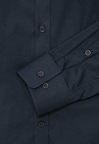 Chemise bleu foncé à boutons, mettant l’accent sur le poignet avec trois boutons et une partie avant avec des boutons visibles et des détails de couture.
