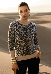 Frau, die in der Wüste steht und einen beigen und schwarzen gemusterten Rollkragenpullover, große goldene Ohrringe und mehrere goldene Armbänder trägt.