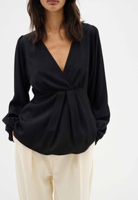 Blusa negra con un profundo escote en V, mangas largas abullonadas y detalles de pliegues en la cintura, combinada con pantalones sueltos de color crema.