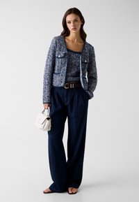 Femme portant une veste en tweed bleu marine et un top, pantalon large bleu marine, ceinture marron, tenant un petit sac à main blanc, debout devant un fond clair uni.