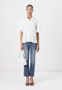 Blusa bianca a maniche corte con un patta frontale testurizzata, abbinata a jeans dritti azzurri e sandali neri con tacco, mentre tiene una piccola borsa bianca.