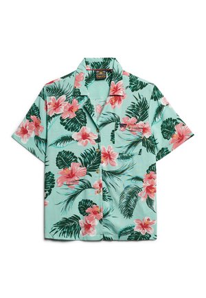 Camicia a maniche corte con bottoni, caratterizzata da uno sfondo azzurro chiaro con fiori di ibisco rosa e foglie verdi; dettaglio della tasca frontale.