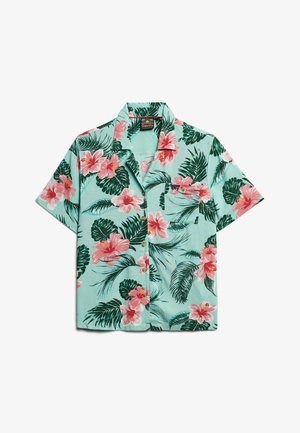 Camisa de manga corta con botones, con fondo azul claro, flores de hibisco rosas y hojas verdes; detalle de bolsillo delantero.