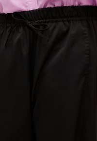 Μαύρο παντελόνι με ελαστική μέση και δεμένο κορδόνι, φορεμένο με ένα ανοιχτό μωβ πουκάμισο που έχει μπει μέσα στο παντελόνι.