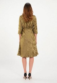 Robe en olive, en soie, avec des manches trois-quarts et un ourlet évasé. Présente une taille froncée et une texture lisse. Portée avec des talons noirs.