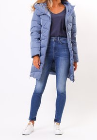 Ljusblå pufferjacka med huva, med dragkedja fram, quiltat mönster och långa ärmar. Buren över en mörk t-shirt och smala jeans.