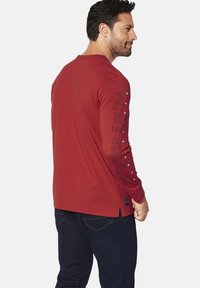 Long-sleeve red T-shirt with black printed text on the left sleeve; features a round neckline and side slit at the hem.