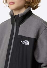 Flis jakna v sivi in črni barvi z visokim ovratnikom. Ima zadrgo in bel logotip na prsih. Mehka tekstura z gladkimi tkaninskimi deli.