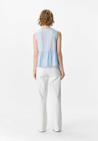 Una mujer con cabello rizado mira hacia un lado, vistiendo una blusa sin mangas de color azul claro y pantalones anchos blancos, frente a un fondo blanco liso.