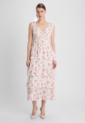 Vrouw draagt een mouwloze witte jurk met roze bloemenprint en gelaagde rok, staand tegen een effen grijze achtergrond.