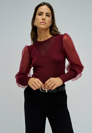 Femme portant un haut bordeaux avec des manches transparentes et bouffantes et un pantalon noir, debout, les mains près d’une boucle de ceinture sur un fond uni.