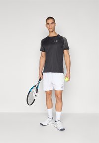 Black athletic t-shirt with textured gray sleeves, paired with white shorts. The model holds a tennis racket and ball, wearing white sneakers.