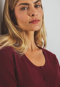 Bordeaux cardigan met een ronde hals, zachte textuur en twee knopen. Heeft een aansluitende fit en wordt gedragen tegen een neutrale achtergrond.