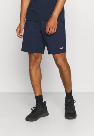 Mężczyzna w granatowych sportowych szortach z białym logo Reebok, czarnych skarpetkach i czarnych sneakersach, stojący na tle jednolitego jasnego tła.