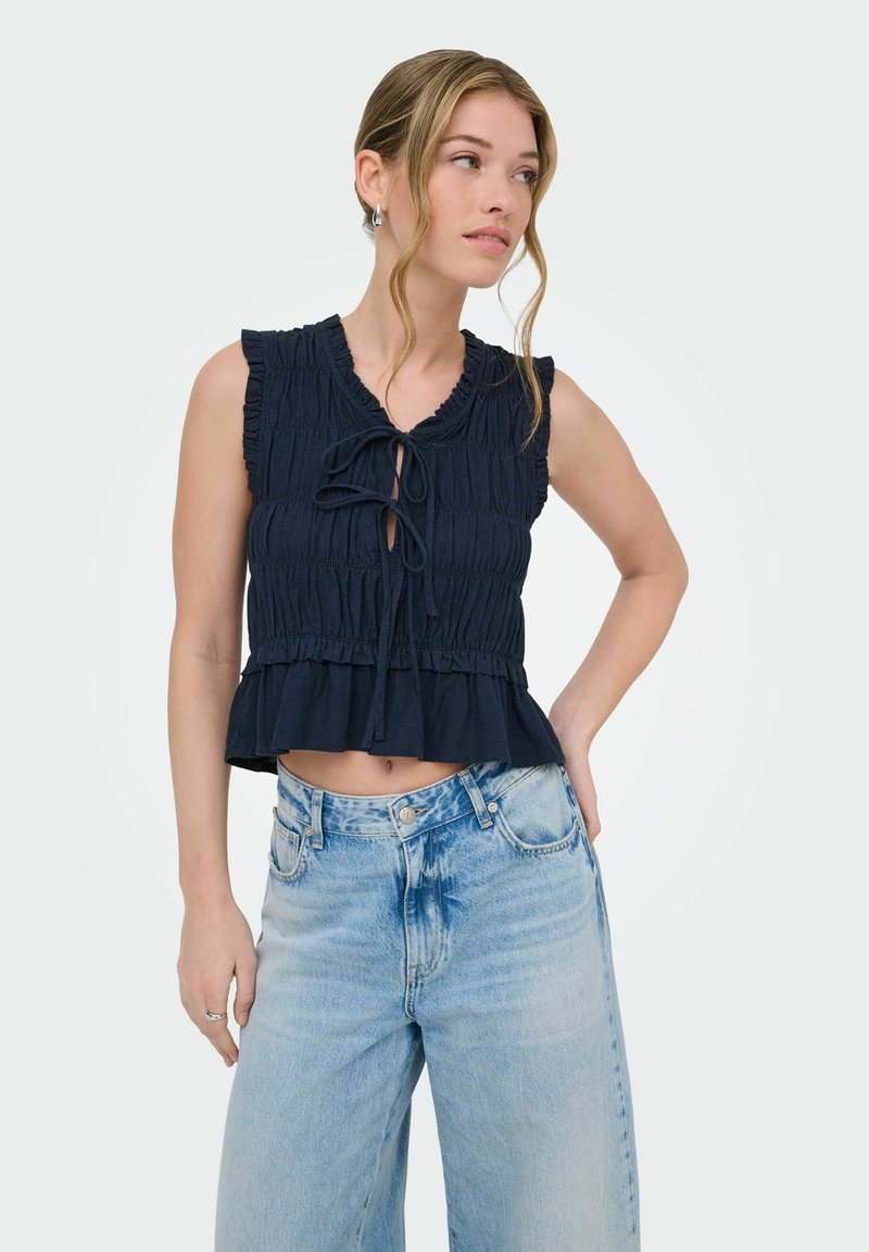 Joven mujer rubia que lleva una blusa negra sin mangas con fruncidos y detalles de lazos, y vaqueros de pierna ancha de color azul claro de talle alto, posando con una mano en la cadera.